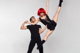 A man wearing all black supports another dancer wearing a red wig as does a tilt jump.